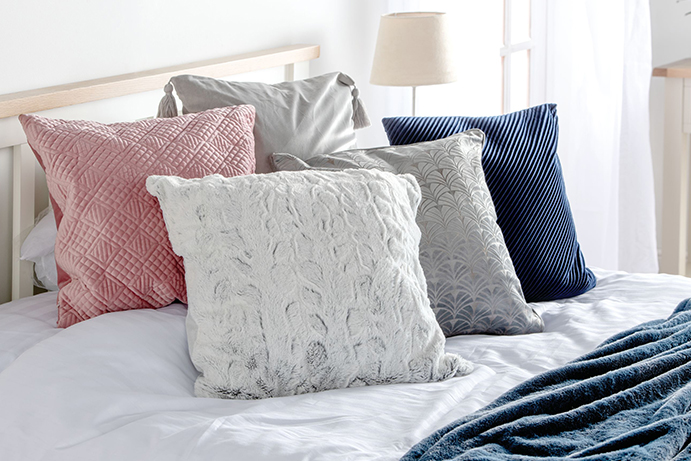 Five cushions in blues, pinks and greys laid on the pillow end of a white double bed
